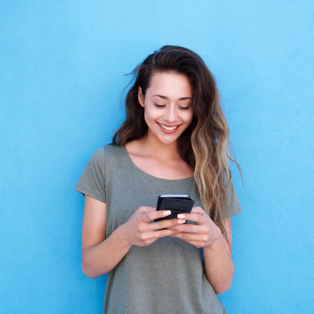 First Fashion PLM Mobile App Person with long wavy hair in gray t-shirt smiling while using smartphone against bright blue background.