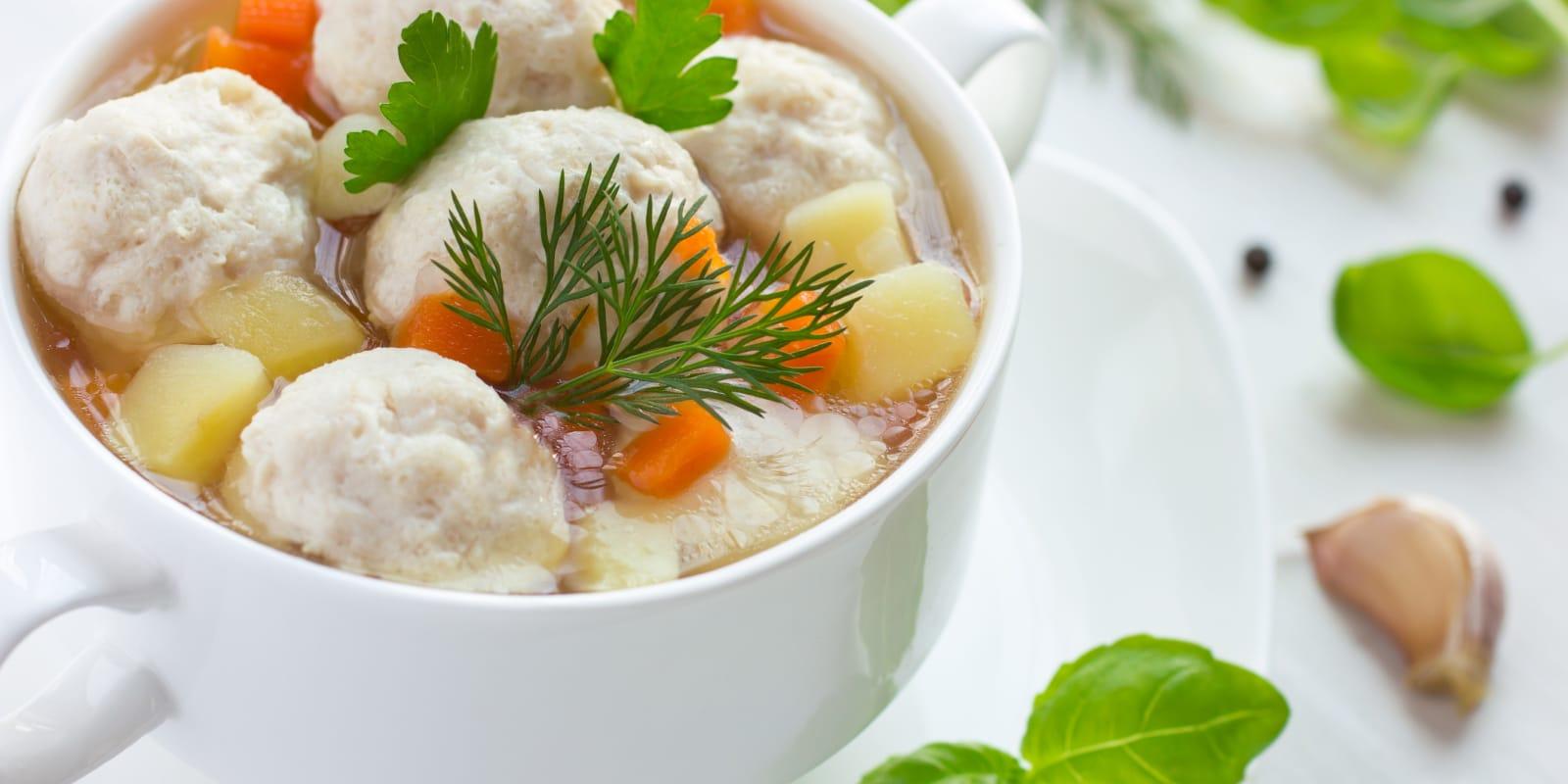 Starpro Transforms R&D Management with Centric PLM Featured A bowl of clear soup with meatballs, diced potatoes, and carrots, garnished with fresh parsley and dill. Perfectly crafted like the innovative solutions from Starpro, it is served in a white bowl on a white surface, adorned by garlic cloves and green leaves.
