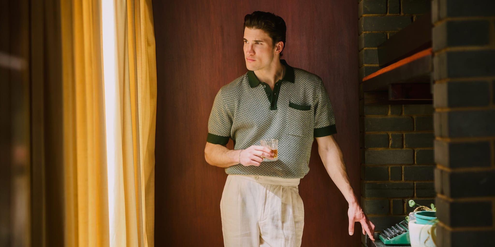 Percival Doubles Product Development Speed and Streamlines Production Workflows with Centric PLM Launch Featured A man in a retro-style short-sleeve shirt and light trousers stands indoors by brickwork and yellow curtains, holding a drink, reflecting thoughtfully on product development as he gazes out the window.
